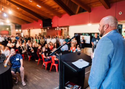 A man in a gray suit speaks at a podium with a microphone to a large, seated audience in a spacious room with wooden ceilings and red walls. The crowd listens attentively.