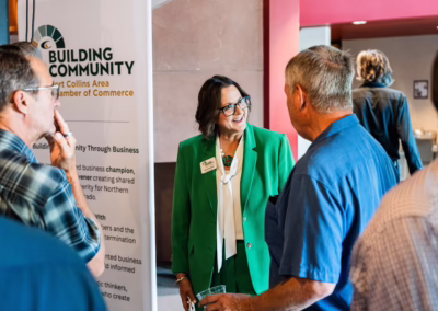A woman in a bright green suit smiles and talks with two men at a business networking event. A banner for the Fort Collins Area Chamber of Commerce stands nearby. Other attendees are visible in the background.