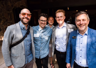 Five men smiling and posing together at an event; some wear name tags and smart-casual attire, standing in a well-lit indoor-outdoor space with stone walls and sunlight.