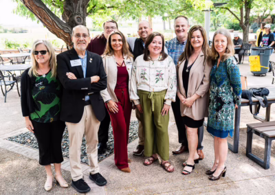 A group of nine adults, dressed in business casual attire, stand together outdoors under a tree, smiling at the camera. A few tables and people are visible in the background on a sunny day.