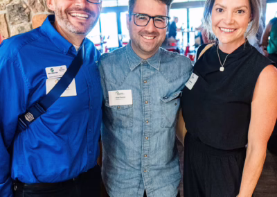 Three people stand smiling indoors at an event. The man on the left wears a bright blue shirt and glasses, the man in the middle wears a denim shirt and glasses, and the woman on the right wears a sleeveless black outfit.