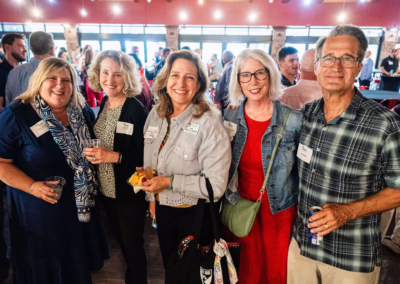 A group of five adults, four women and one man, smile for a photo at an indoor social event. They are wearing name tags and casual clothes, with people and tables visible in the background.