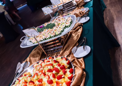 A buffet table covered with a dark green tablecloth and gold runner, featuring large trays of fresh fruit and assorted foods, small plates, tongs, and a vase of white flowers in a decorated indoor setting.