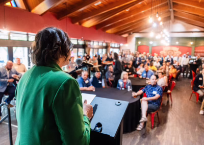 A person in a green jacket speaks at a podium to a large seated audience in a spacious, well-lit room with wooden ceilings and red walls. The crowd listens attentively, some seated at round tables.