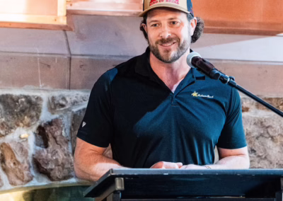 A man in a black shirt and Budweiser cap speaks at a podium with a microphone. The podium features a sign that reads "Building Community Fort Collins Area Chamber of Commerce.