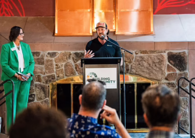 A man wearing a cap speaks at a podium labeled "Building Community" while a woman in a green suit stands to his left, and audience members listen in the foreground. Stone wall and copper fixtures are in the background.