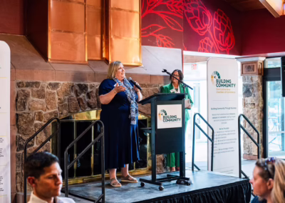 Two women stand on a small stage speaking at a podium with a "Building Community" sign, inside a venue with stone walls and red decor. Audience members listen in the foreground.