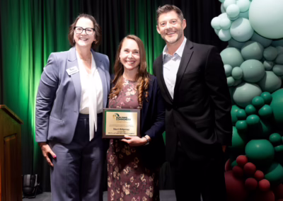 Three people stand smiling in front of green and teal balloon decorations. The woman in the center holds an award plaque. The woman on the left wears a gray suit, and the man on the right wears a black suit.
