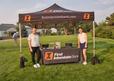 Two women stand smiling under a First Interstate Bank tent at a golf course, with a table displaying promotional materials and a laptop on the grass.