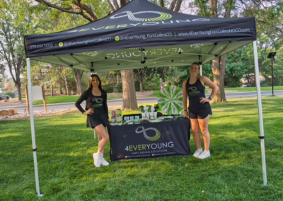 Two women in black outfits stand by a “4EverYoung” anti-aging solutions booth under a branded canopy, with products displayed on a table, set in a sunny, tree-filled park.
