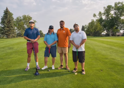 Four men stand on a golf course with clubs, posing for a photo on green grass with trees and a cloudy sky in the background.