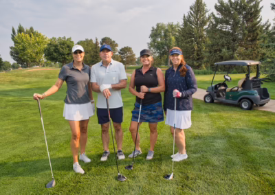 Four people stand on a golf course holding clubs, dressed in golf attire, with a golf cart and trees in the background.