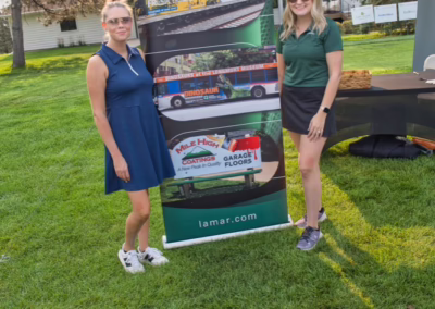 Two women stand smiling beside a vertical Lamar advertising banner at an outdoor event, with green grass, trees, and a tent canopy in the background.