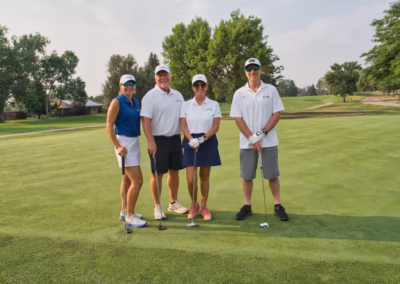 Four people stand on a golf course green, holding clubs and smiling. They wear golf attire, including visors and sunglasses. Trees and a clear sky are visible in the background.