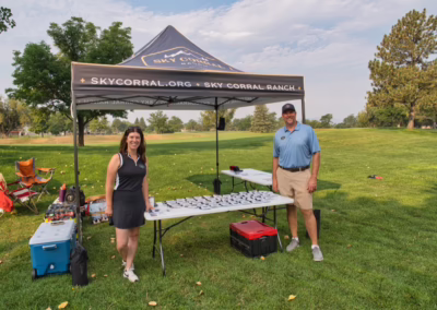 A woman and man stand under a canopy labeled “Sky Corral Ranch” next to a table with plates on a grassy field, with chairs and trees in the background on a sunny day.
