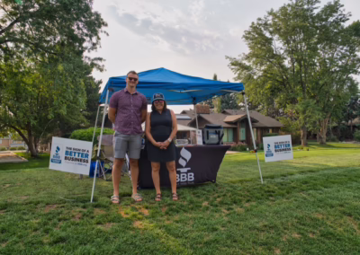 Two people stand smiling in front of a blue tent with a BBB table and signs reading “This sign of a better business.” The scene is set on grass near trees and a house on a sunny day.