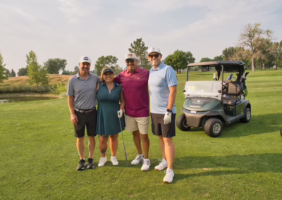 Four people in golf attire stand together smiling on a golf course near a golf cart, with trees and grass in the background on a sunny day.