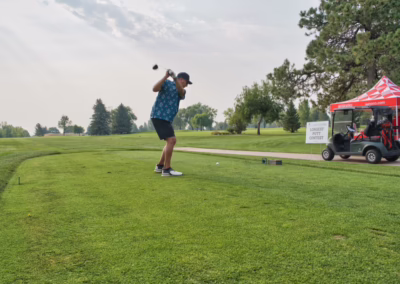 A man swings a golf club on a green course near a red tent and a golf cart, with trees and a cloudy sky in the background.