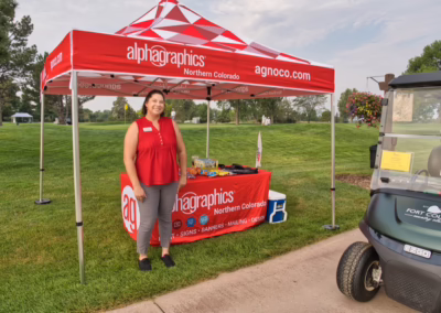A woman stands smiling under a red Alphagraphics-branded tent with snacks on a table, on a golf course next to a golf cart, with green grass and trees in the background.