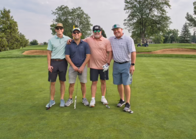 Four men stand smiling on a golf course green, wearing casual golf attire and caps. Two hold golf clubs. Trees, a sand trap, and a golf cart are visible in the background.