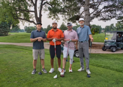 Four men stand on a golf course with clubs, smiling at the camera. Golf carts and trees are in the background. They are dressed in casual golf attire.