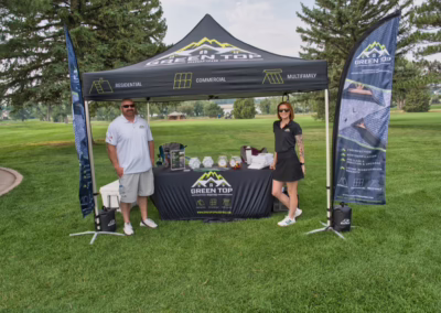 Two people stand in front of a promotional tent for Green Top Roofing on a grassy area, with banners, a table of items, and trees in the background.