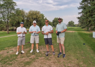 Four men wearing golf attire stand on a golf course, smiling and holding clubs, with trees and a cloudy sky in the background.
