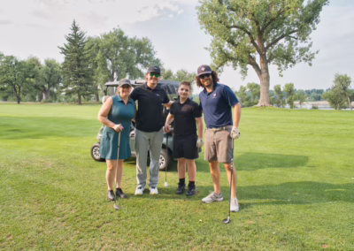 Four people stand smiling on a golf course with a golf cart behind them. They each hold golf clubs and wear golf attire. Trees and a lake are visible in the background under a partly cloudy sky.