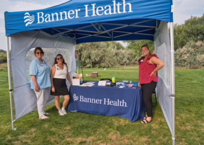 Three women stand at a Banner Health booth outdoors, under a blue canopy on a grassy field. The table displays informational materials and promo items with trees visible in the background.