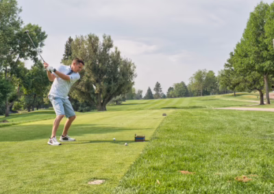 A person in a white shirt and shorts swings a golf club on a green golf course, preparing to hit a golf ball. Trees and grass surround the area under a partly cloudy sky.