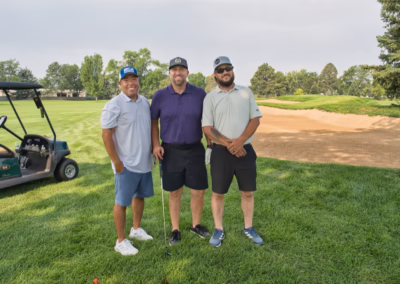 Three men pose together on a golf course near a sand bunker, smiling at the camera. A golf cart is parked on the grass beside them. Trees and greenery are visible in the background.