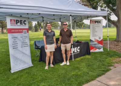 Two people stand smiling under a tent at a PEC booth set up on a grassy field, with banners and a table displaying information about professional engineering services. Trees and golfers are visible in the background.