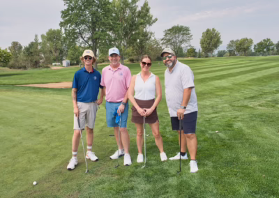 Four people stand on a green golf course with clubs, smiling at the camera. Trees and sand traps are visible in the background under a cloudy sky.