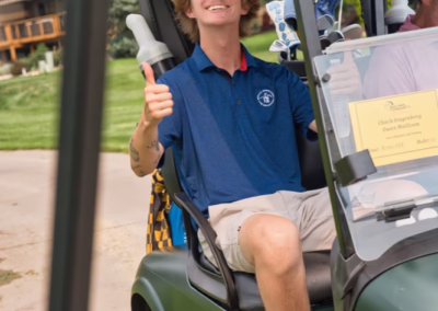 A young man in a blue polo shirt and cap sits in a golf cart, smiling and giving a thumbs-up. He wears sunglasses and sports shoes, with a golf bag visible behind him on a sunny day.