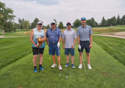 Four people stand on a green golf course, each holding golf clubs. One person holds a plush toy. They are dressed in casual golf attire, sunglasses, and hats, posing for a group photo under a cloudy sky.