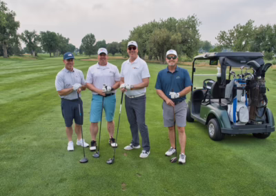 Four men stand on a golf course with clubs and gloves, posing beside a golf cart on a green fairway lined with trees under a cloudy sky.