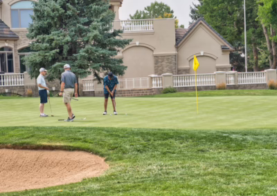 Three people stand on a golf green, one preparing to putt near the hole marked by a yellow flag. A large house and trees are in the background. A sand bunker is visible in the foreground.