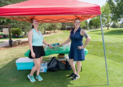 Two women stand under a red tent on a grassy area, smiling at the camera. They are holding a small black sign that reads "ENTER TO WIN." There are coolers, beverages, and supplies on the table between them. Trees and a path are visible in the background.