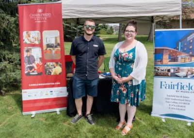 Two individuals are standing outside under a canopy, next to banners promoting Candlewood Suites and Fairfield by Marriott. The person on the left is wearing sunglasses and a dark outfit, while the person on the right is dressed in a green floral dress and a white cardigan.