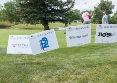 Two men are on a golf course. One man is in mid-swing, while the other watches. Several sponsor signs are displayed on the grass, including Tetrad Real Estate, PCL Construction, Banner Health, and Noosa Yoghurt. Trees and a pond are visible in the background.