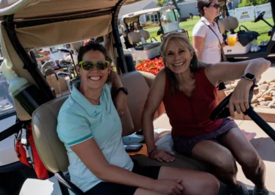 Two women are sitting on a golf cart, smiling brightly. One woman is wearing sunglasses and a light blue polo shirt, while the other is in a sleeveless top and shorts. They appear to be enjoying a sunny day at the golf course with other people and carts in the background.