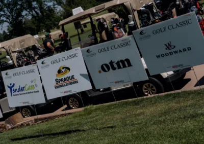 A row of golf carts is parked on a path with several sponsored signs in front of them that read, "Golf Classic." Sponsors include TenderCare, Sprague Roofing and Exteriors, OTM, and Woodward. The event appears to be set on a sunny day at a golf course.