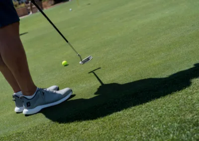 A person in gray golf shoes prepares to putt a yellow golf ball on a green course, their shadow cast on the grass.