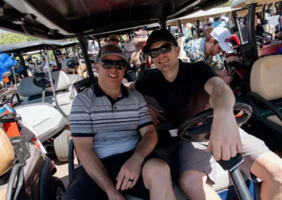 Two men sitting in a golf cart, both wearing sunglasses and caps. The man on the left is older in a striped shirt, and the man on the right is younger in a black shirt. Other golf carts and people are visible in the background, suggesting a busy golf course.