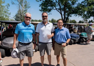 Three men are standing and smiling on a sunny day at a golf course, dressed in golf attire. Behind them are several golf carts and other people. Trees and a clear blue sky are visible in the background.