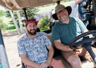 Two men are sitting in a golf cart, smiling at the camera on a sunny day. The man on the left wears a floral shirt and red cap, while the man on the right has a beard, glasses, a wide-brim hat, and a teal shirt. They appear to be on a golf course with carts and clubs in the background.