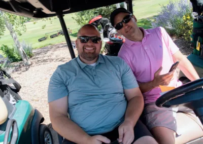 Two men sitting in a golf cart on a sunny day. Both are wearing sunglasses and smiling at the camera. The man on the left is in a light blue polo and khaki shorts, while the man on the right is in a pink polo and beige shorts, holding a smartphone. Golf equipment is visible in the background.