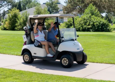 Two people riding in a golf cart on a sunny day. The person on the passenger side is waving and smiling, wearing sunglasses and a striped shirt. The driver is wearing a hat and blue shirt. They're on a paved path with lush green trees and grass in the background.