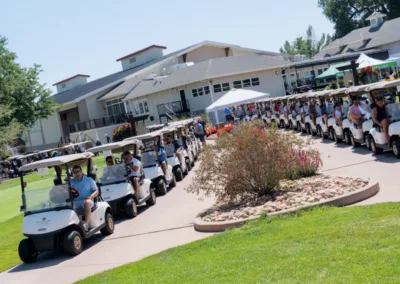 A long line of golf carts filled with people wearing casual attire are parked on a paved path outside a large clubhouse with a green lawn and landscaped bushes nearby, on a sunny day. The scene indicates a social or organized golf event.