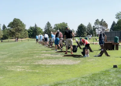 A group of people practicing on a golf driving range, each at individual stations. Some are swinging clubs, while others are setting up their shots. The range is surrounded by trees under a clear blue sky.